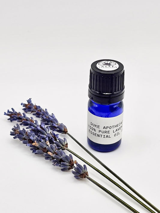 A small blue bottle of lavender essential oil with a black cap, labeled 'Lavender Pure Essential Oil'. Next to the bottle, there are hand-harvested lavender flowers spread out on a white background.