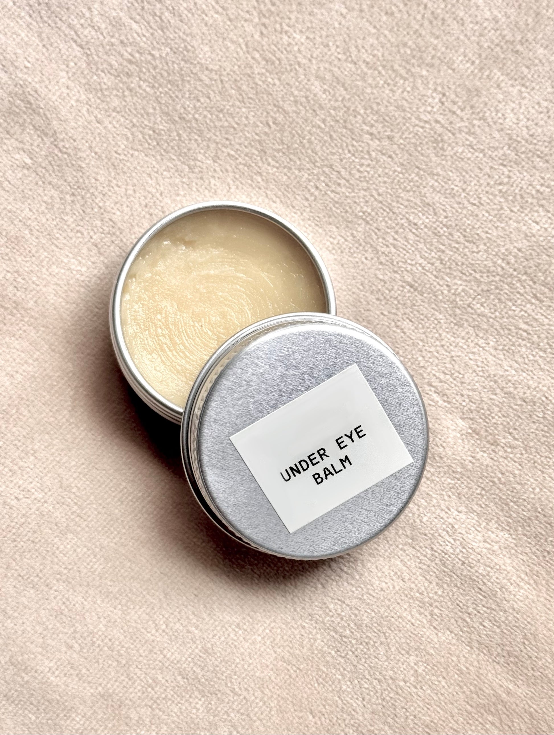 Open tin of under eye balm with a label on a beige background