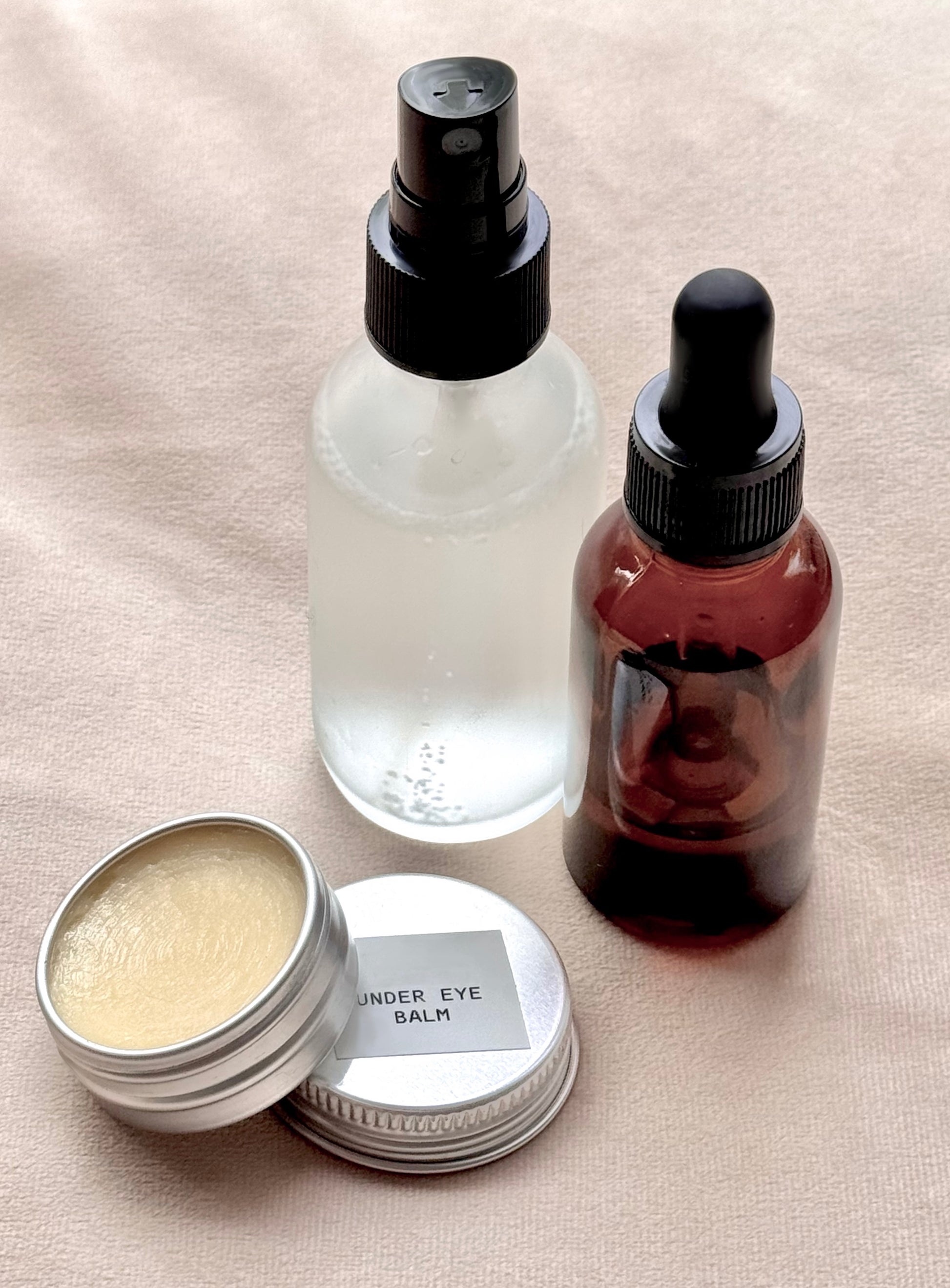 Skincare products including a jar of under eye balm, a spray bottle, and a dropper bottle on a beige surface.