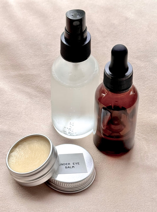 Skincare products including a jar of under eye balm, a spray bottle, and a dropper bottle on a beige surface.