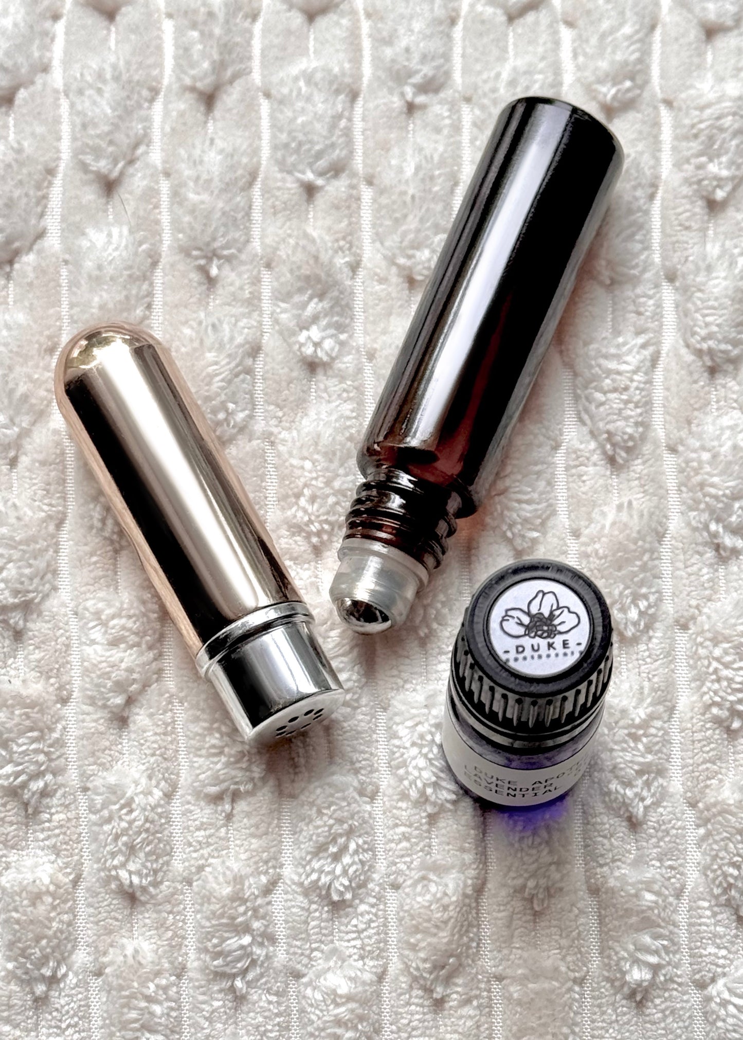 Two silver rollerball bottles and a small round bottle with a visible brand logo on a textured white surface.