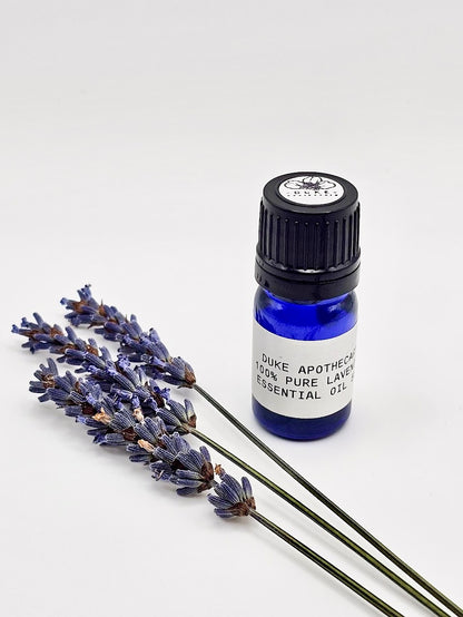 A small blue bottle of lavender essential oil with a black cap, labeled 'Lavender Pure Essential Oil'. Next to the bottle, there are hand-harvested lavender flowers spread out on a white background.