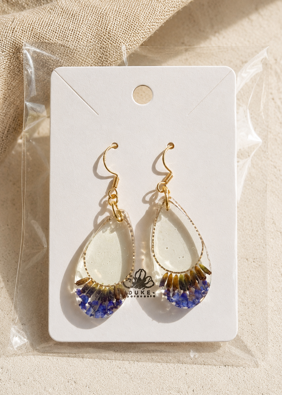 Teardrop-shaped earrings with gold and blue accents on a white card with 'DUKE' branding, placed on a beige fabric background.