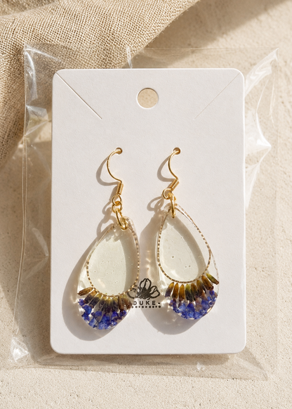 Teardrop-shaped earrings with gold and blue accents on a white card with 'DUKE' branding, placed on a beige fabric background.