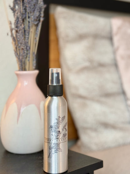 Deep Sleep linen & room spray sitting on a side table in front of a vase with dried lavender in it next to a bed with lots of soft pillows 