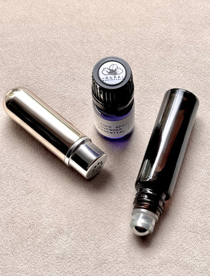 Three small containers on a beige surface, including a silver tube, a small bottle with a black cap, and a black rollerball.