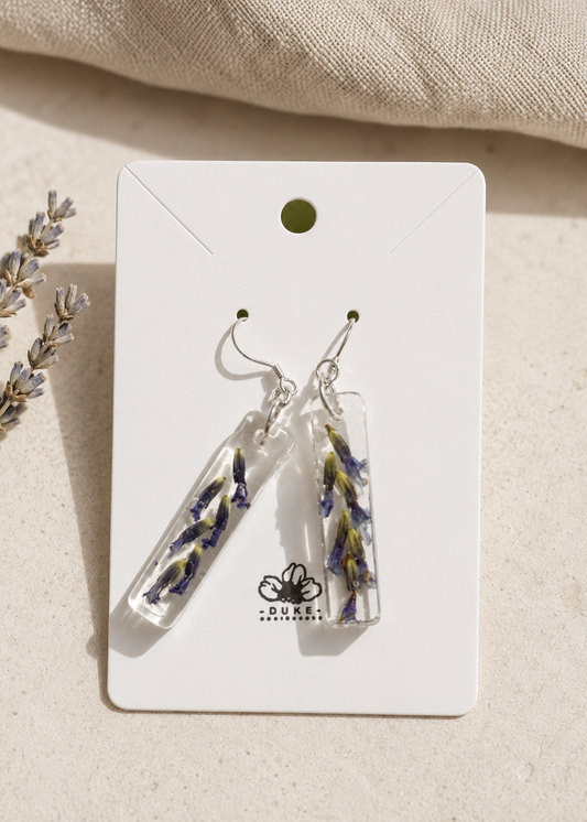 Clear earrings with floral designs on a Duke & Company branded card, placed on a beige fabric background.